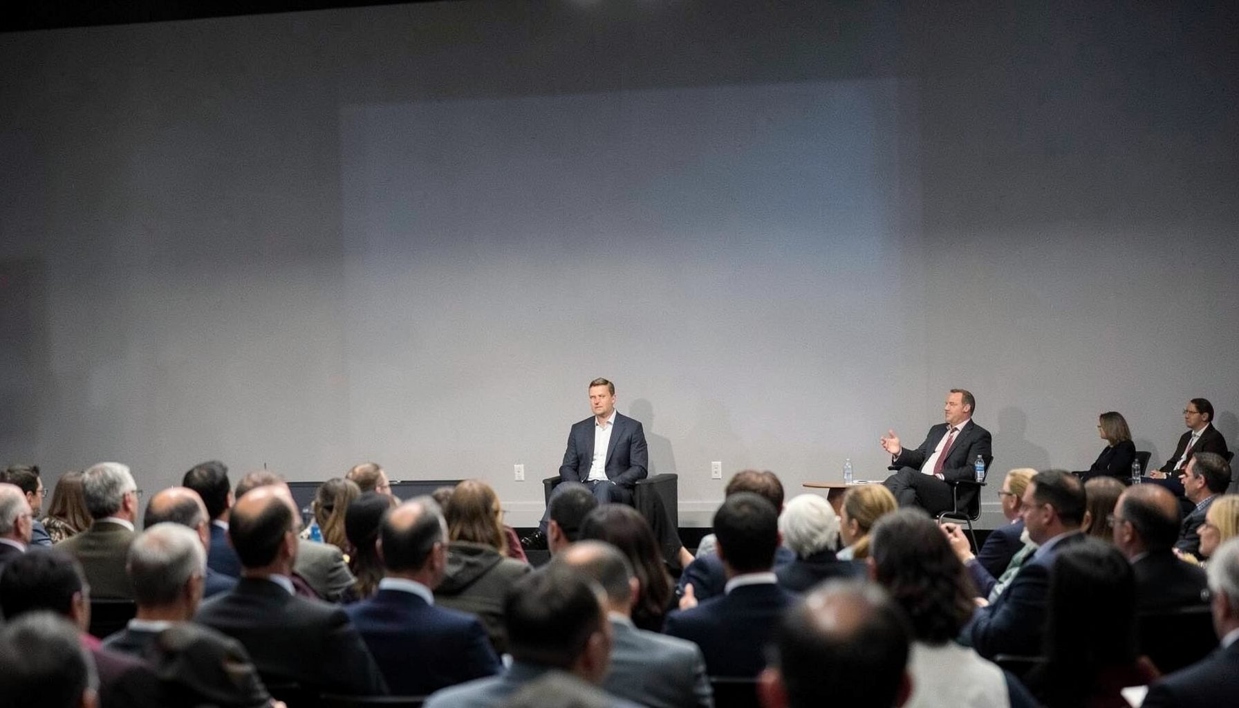 Presenter speaking at a broadcast industry panel discussion, neutral grey stage with projection screen and seated audience